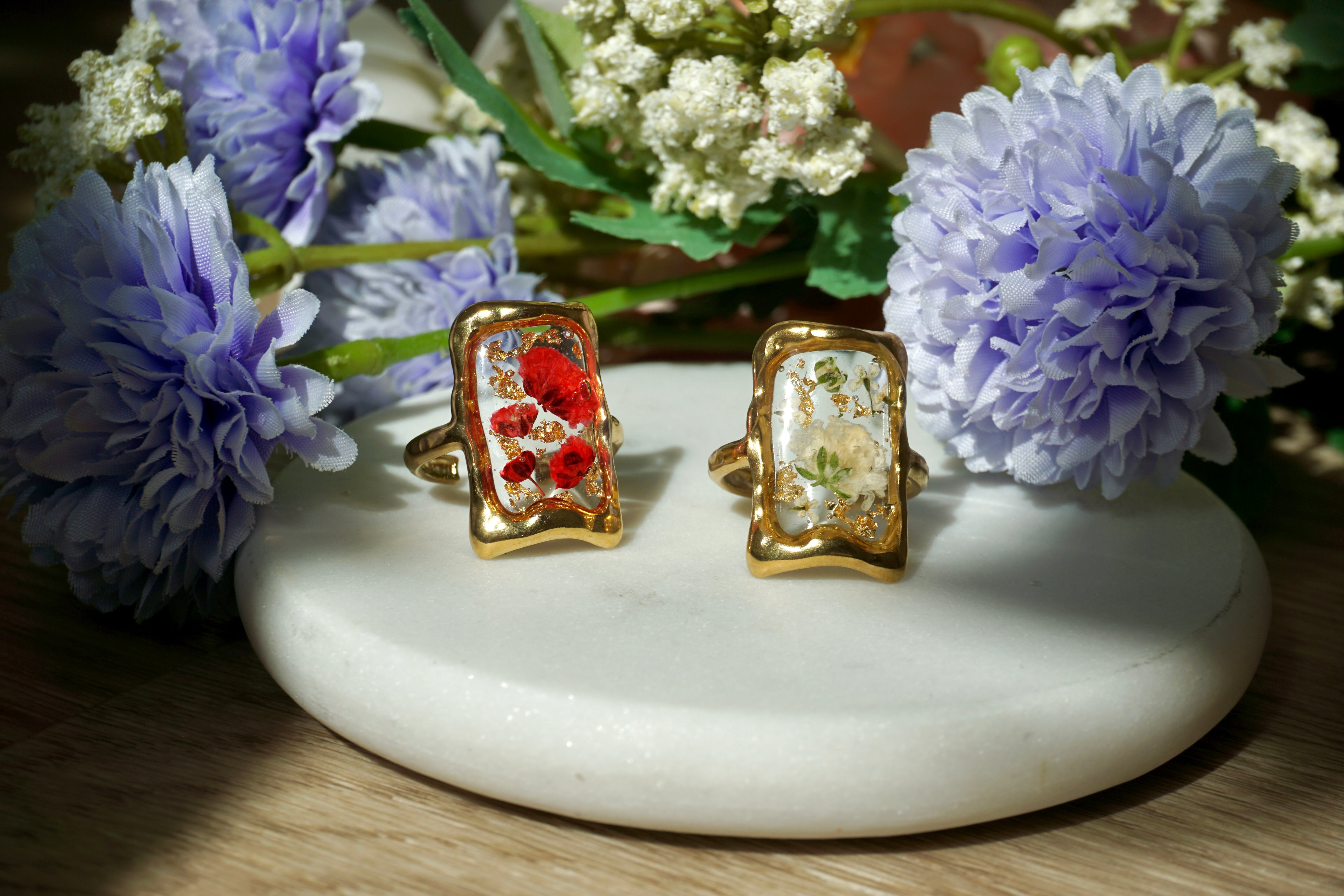 Two gold rings with floral designs on a white stone surface with purple flowers.