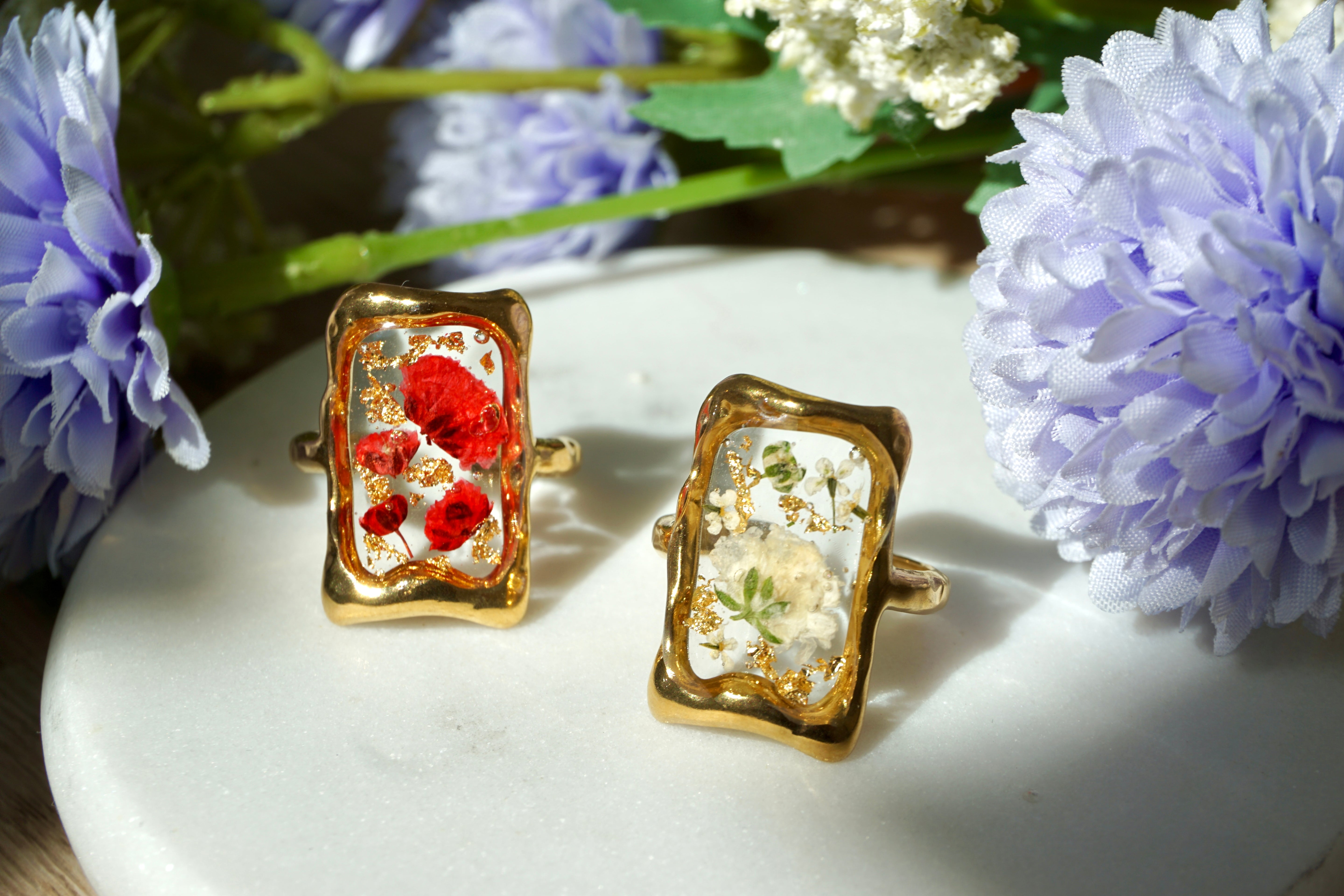 Gold-framed rings with floral designs on a white surface surrounded by flowers.