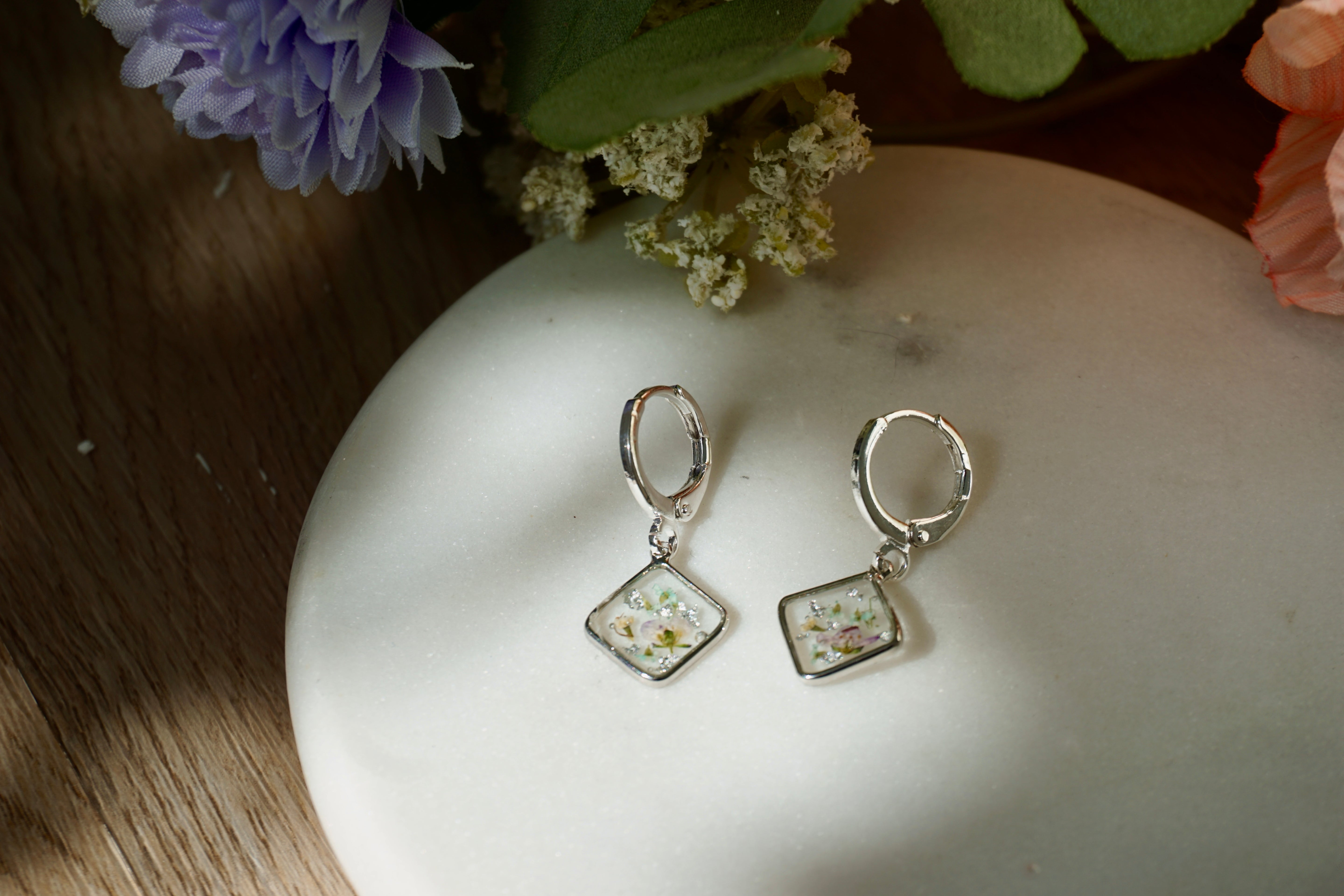 Silver earrings with square designs on a white surface with flowers in the background