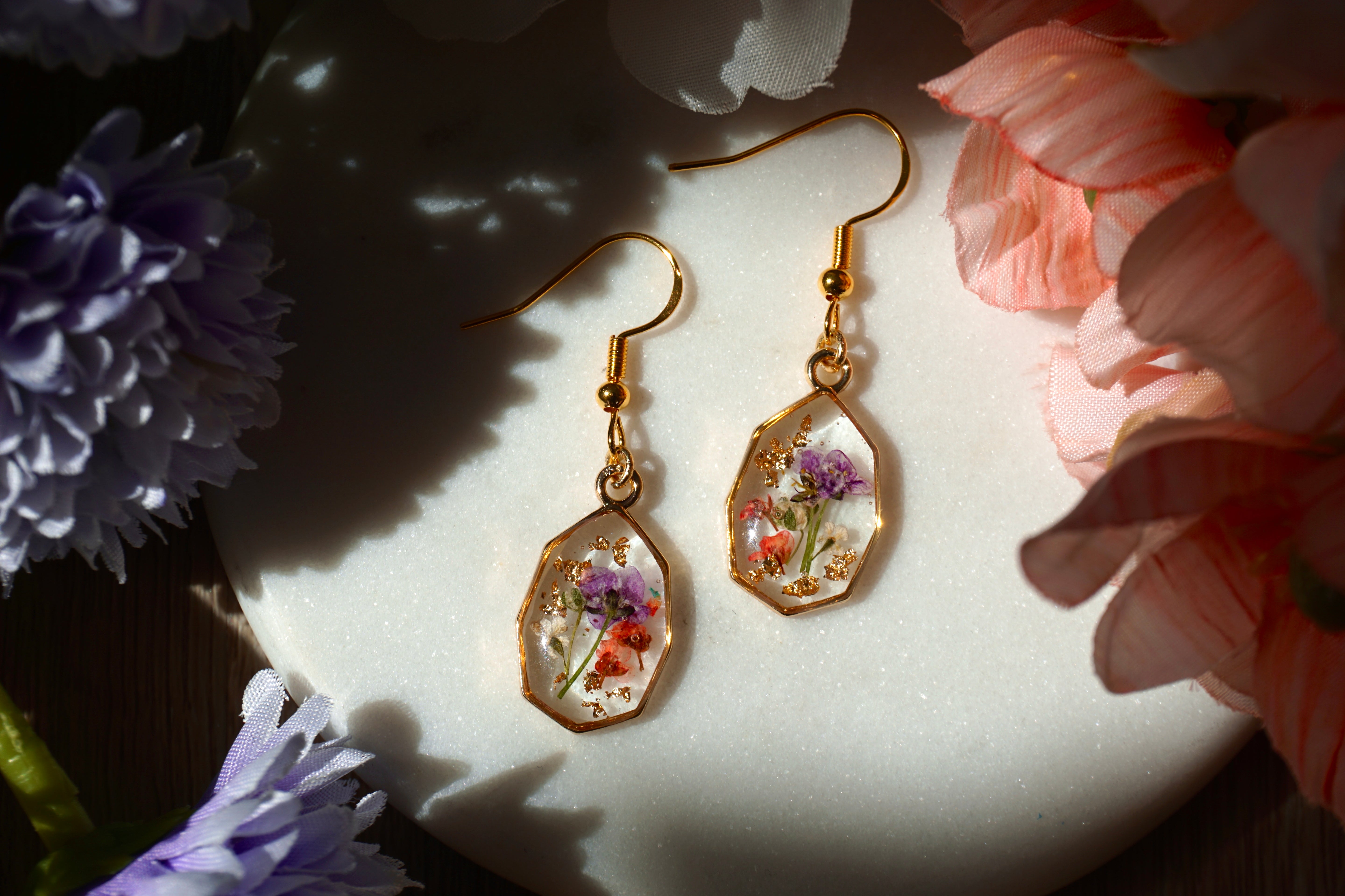 Gold earrings with floral designs on a white surface surrounded by flowers