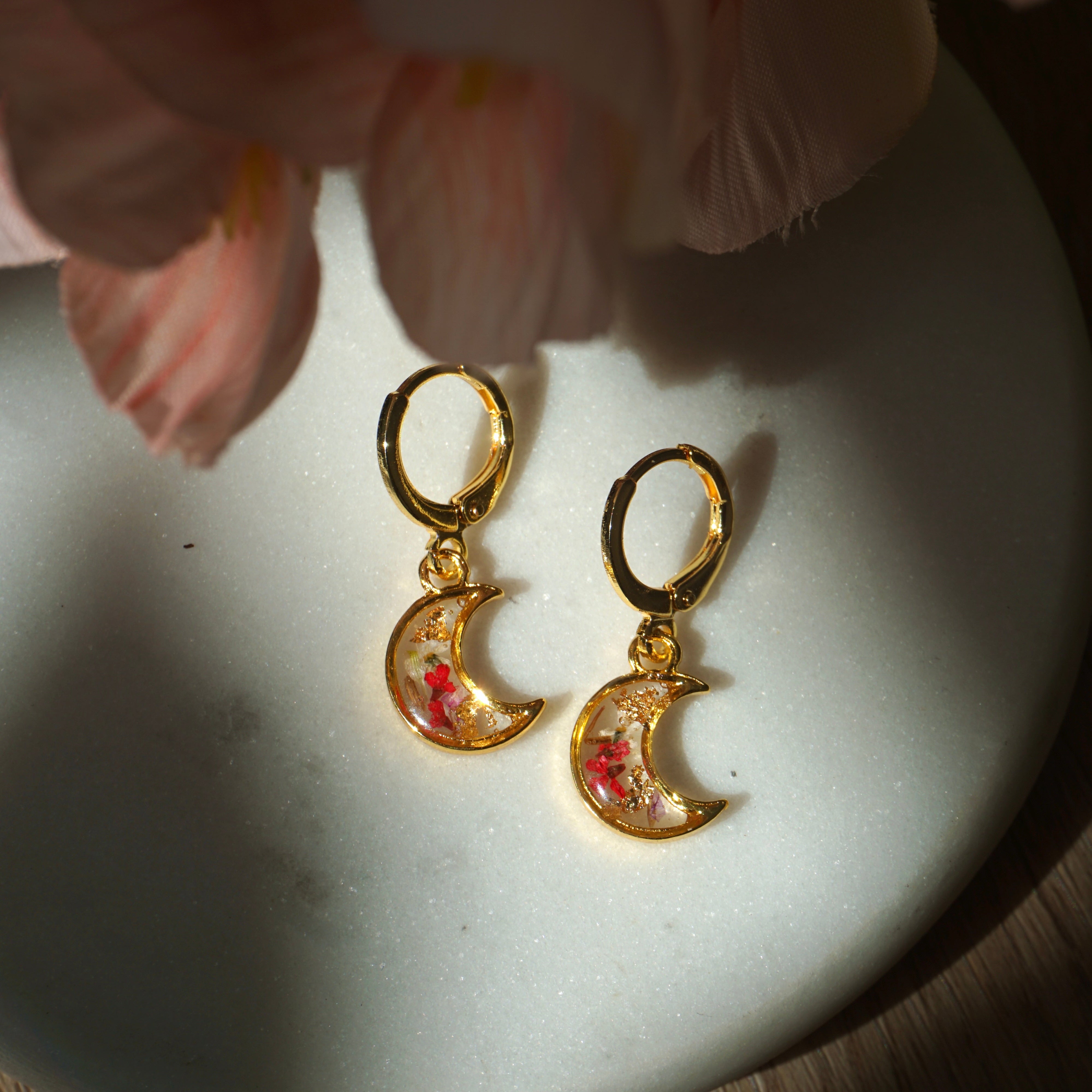 Gold moon-shaped earrings with floral details on a white dish, with a soft pink flower in the background.