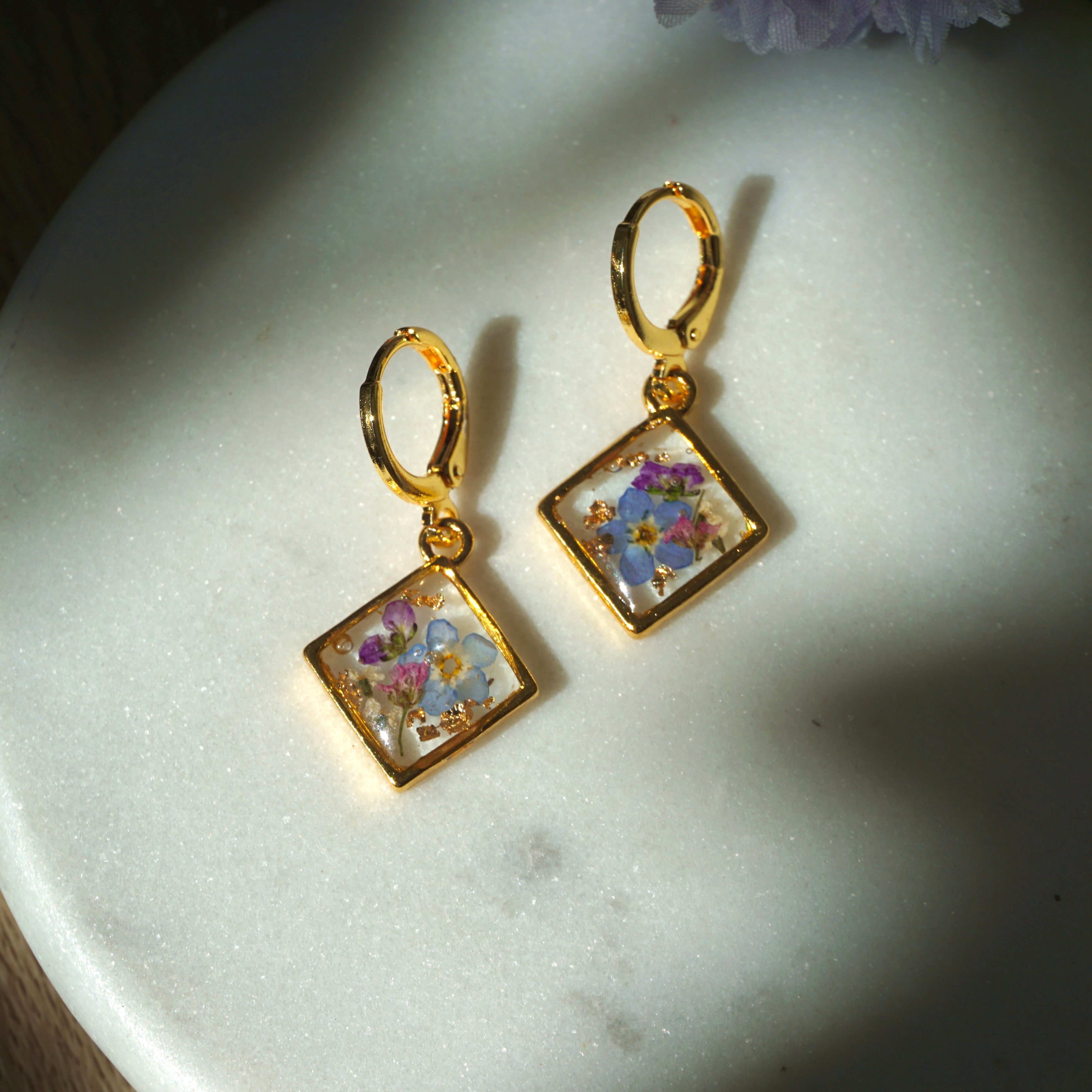 Gold earrings with floral designs in square frames on a marble surface.