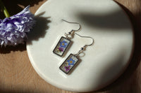 Pair of earrings with floral designs on a white plate with a purple flower beside it.