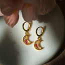 Gold moon-shaped earrings with floral details on a white dish, with a soft pink flower in the background.