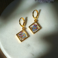 Gold earrings with floral designs in square frames on a marble surface.